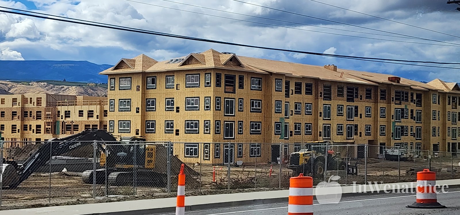 Wide view of a wood-framed multi-story building under construction on a dry grassy site, with stacked materials in front and hills under a cloudy sky behind it.