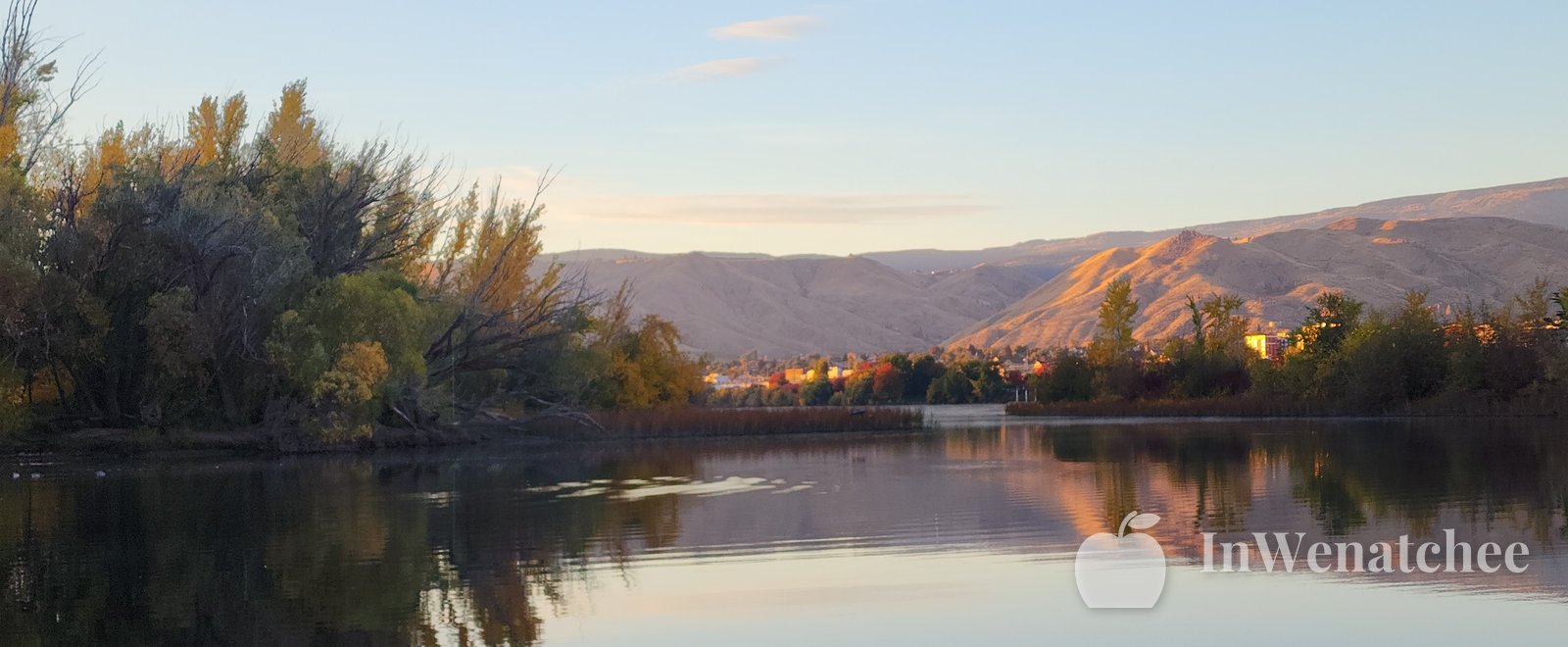 Calm Columbia River at sunset from the Wenatchee riverfront, with a pale sky, a low hill on the horizon, and reeds along the near shore.