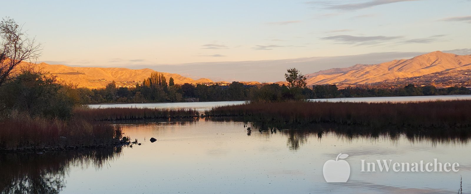 Wide sunset over the Columbia River at the Wenatchee riverfront, with a bright sky, rolling hills, and light reflected across the water.