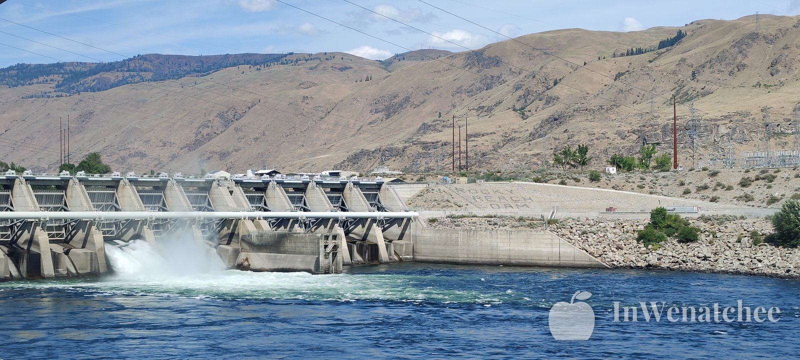 Water spilling from the Rocky Reach Dam spillway into the blue Columbia River, with a dry hillside behind.