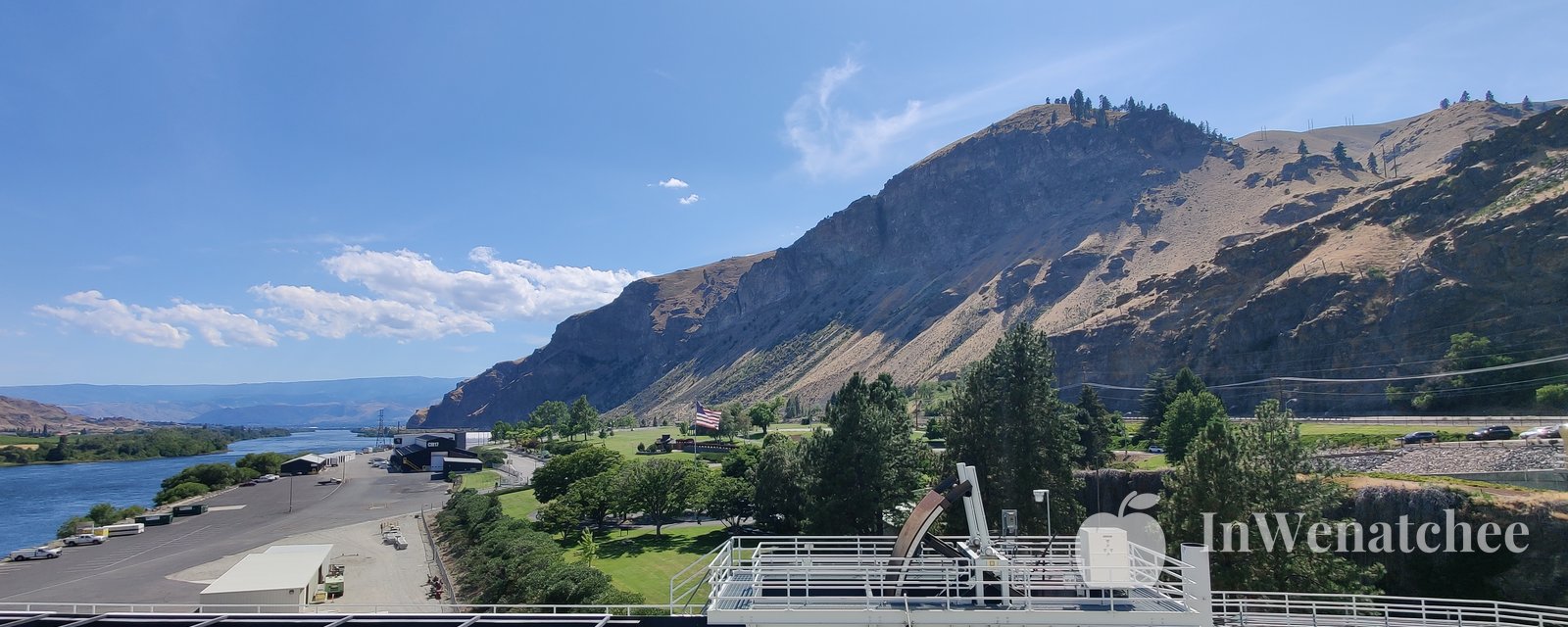 View over the Rocky Reach Dam powerhouse area, with the Columbia River on the left and a steep, rocky hillside rising behind green lawns and trees.