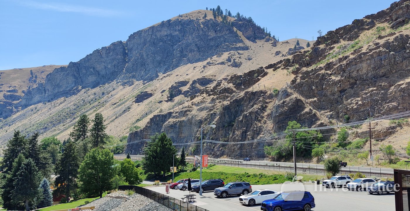 A steep, rocky hillside above the Rocky Reach parking area, with evergreen trees and parked cars below and blue sky above.