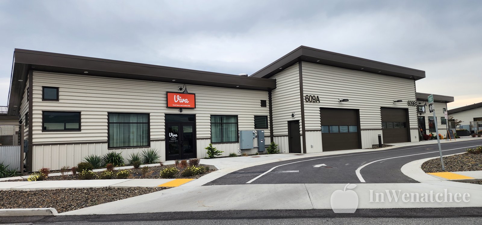 Modern light-colored building with a red Viva sign and garage doors on a new street under a cloudy sky.